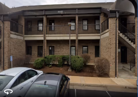 Two-story brick building with balconies, shrubs, and parked cars along the front.