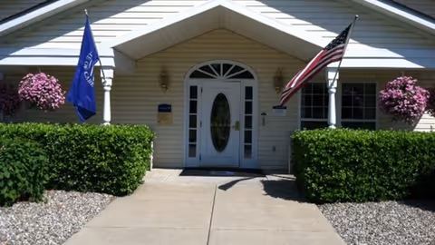 Front entrance of a single-story building with a white door featuring an oval glass window. There are two flags on either side of the entrance, one American flag and one blue flag. The entrance is flanked by green bushes and hanging baskets with pink flowers. The building has light-colored siding and a peaked roof above the door.