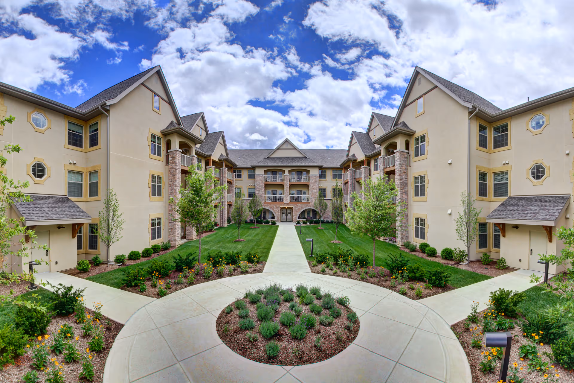 Exterior view of The Plaza at The Barrington of Carmel showing a landscaped courtyard with green grass, small trees, and flower beds surrounded by a three-story beige building with balconies and multiple windows under a partly cloudy blue sky.