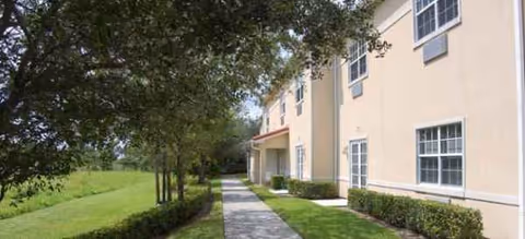 Beige senior living building with a walkway, lawn, trees, and hedges.