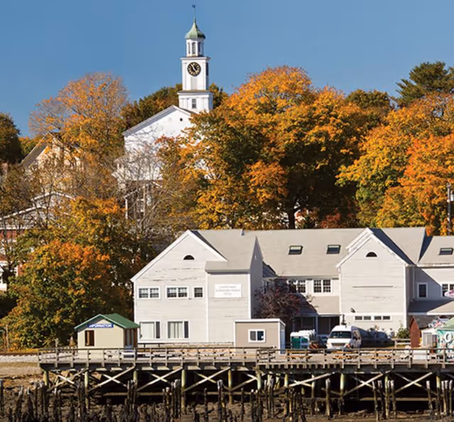 Exterior waterfront building with a clock tower, dock, and autumn trees in the background.