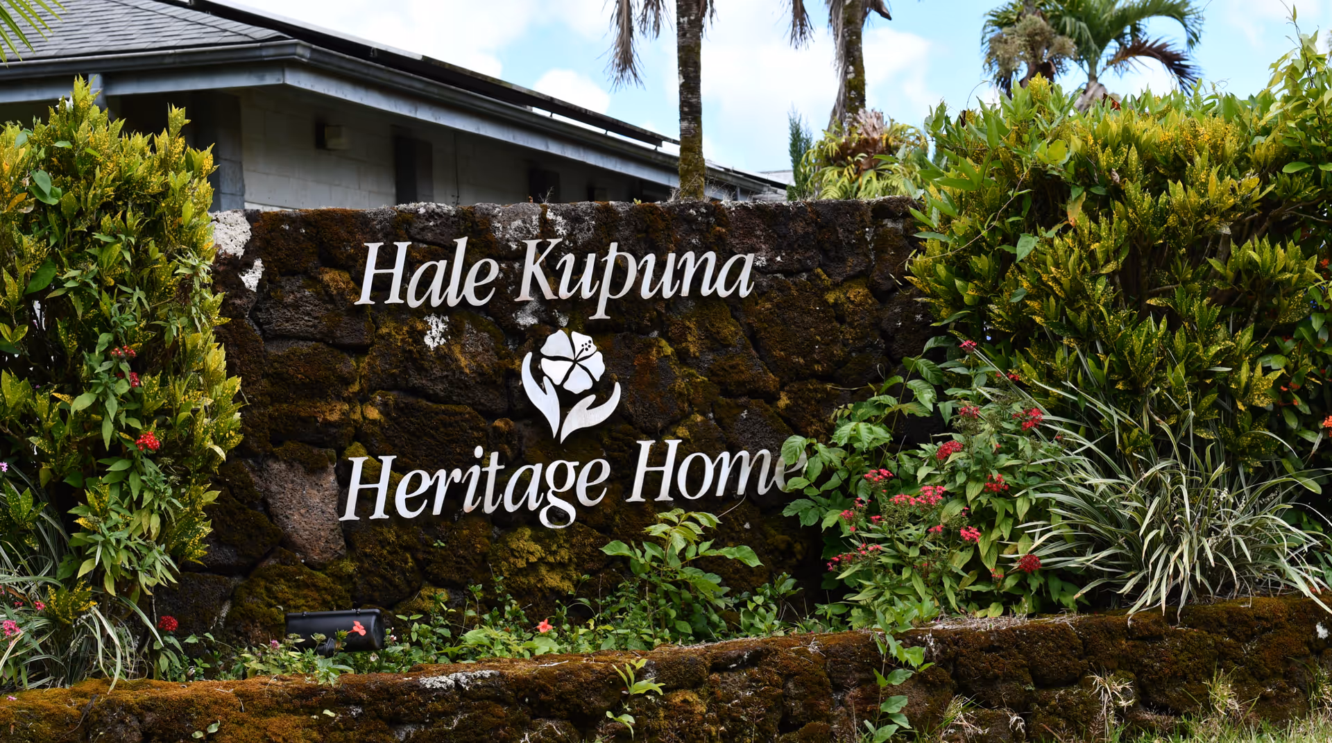 Exterior rock wall with Hale Kupuna Heritage Home sign surrounded by tropical plants.