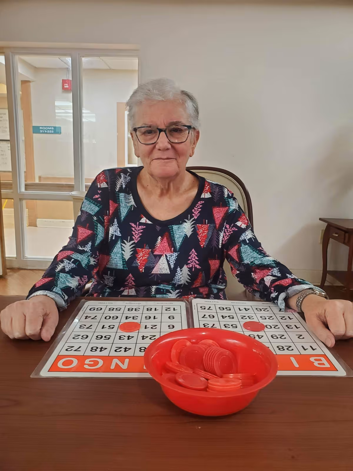 An elderly woman with short gray hair and glasses sits at a table playing bingo. She is wearing a colorful patterned shirt and has two bingo cards in front of her with red markers on some numbers. A red bowl filled with red bingo chips is also on the table. The background shows a hallway with a sign indicating rooms 311-322.