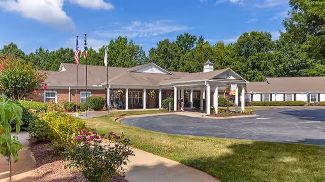 Front entrance of a single-story brick senior living facility with a covered porte-cochère, flagpoles, and landscaped grounds under a blue sky.