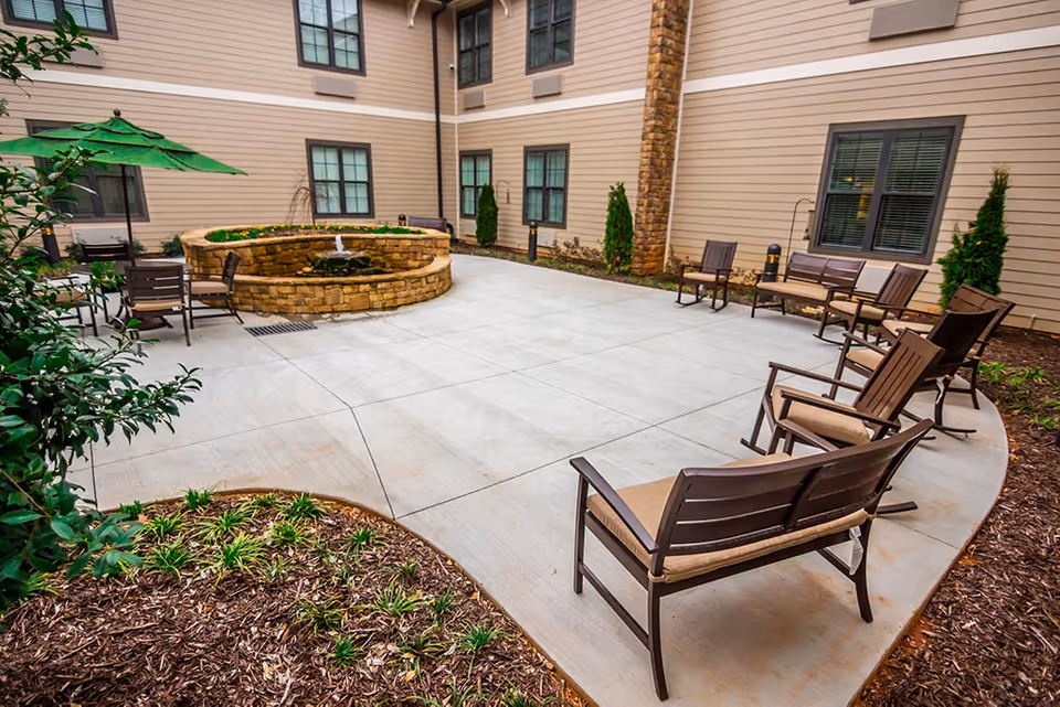 Outdoor courtyard area at Brickmont at Acworth featuring a circular stone water fountain in the center, surrounded by several cushioned chairs and benches arranged on a concrete patio. The courtyard is bordered by garden beds with mulch and plants, and the building exterior has beige siding with multiple windows.