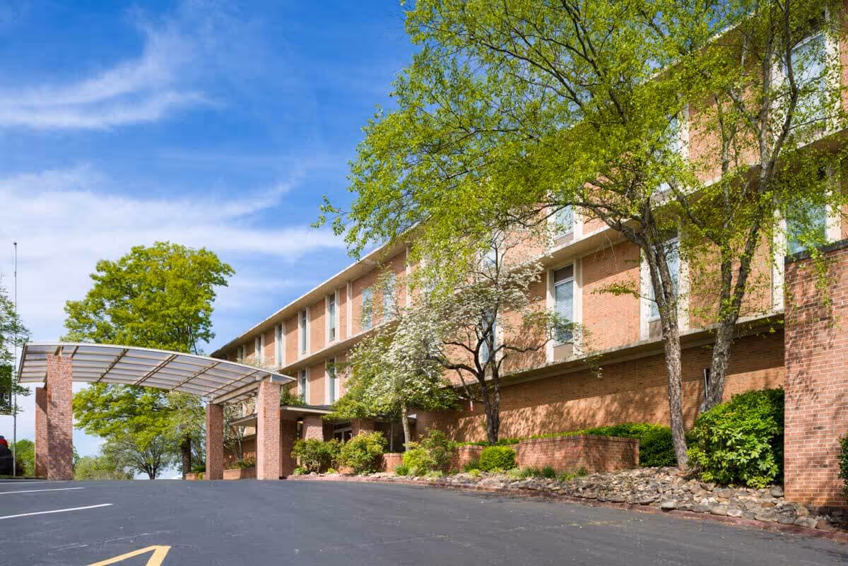 Brick three-story senior living building entrance with a covered porte-cochere, trees, and a paved driveway.