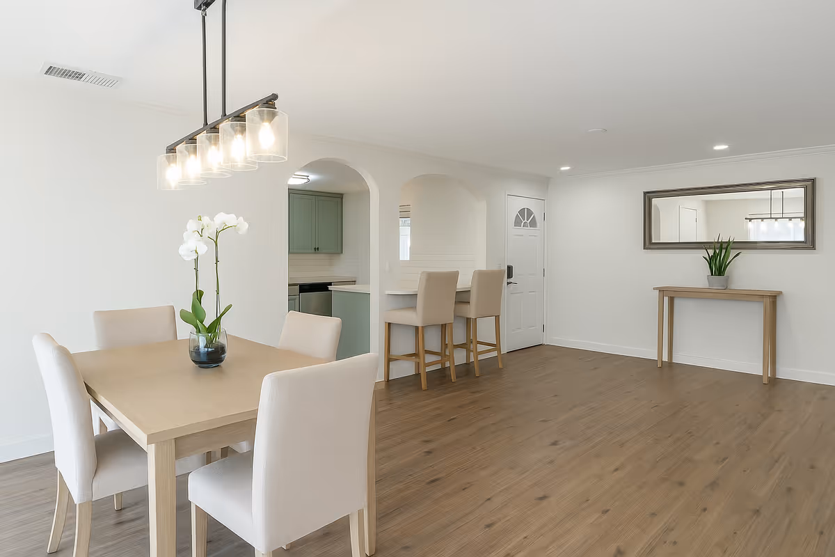 Open dining area with a wooden table, beige chairs, and a kitchen arch in background.