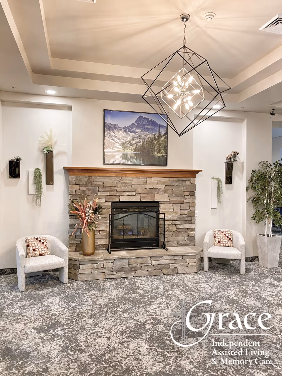 A lodge-style living area with a stone fireplace, two white chairs, plants, and a geometric chandelier.