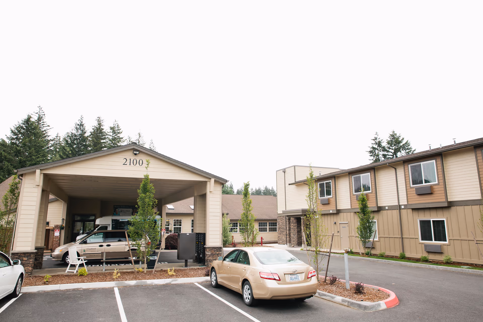 Exterior view of Village Concepts of Chehalis - Woodland Village showing a covered entrance with the number 2100 above it, a parking lot with several cars, and a two-story building with beige and brown siding surrounded by trees.