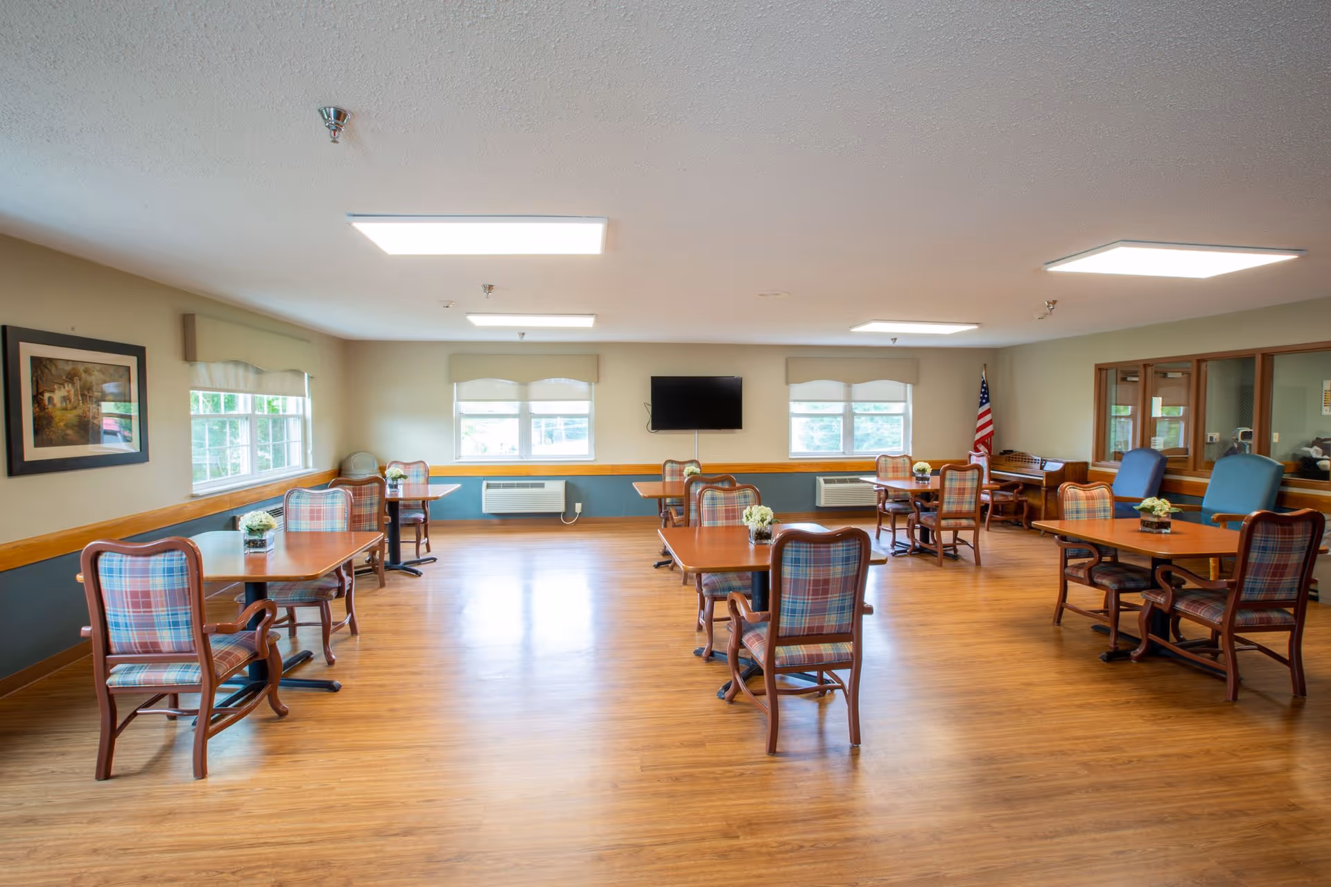 A spacious dining room with wooden floors and several wooden tables surrounded by plaid upholstered chairs. The room has large windows with beige valances, a wall-mounted flat screen TV, an American flag in the corner, and a piano near the back wall. The walls are painted beige with a blue lower section and decorated with a framed picture.
