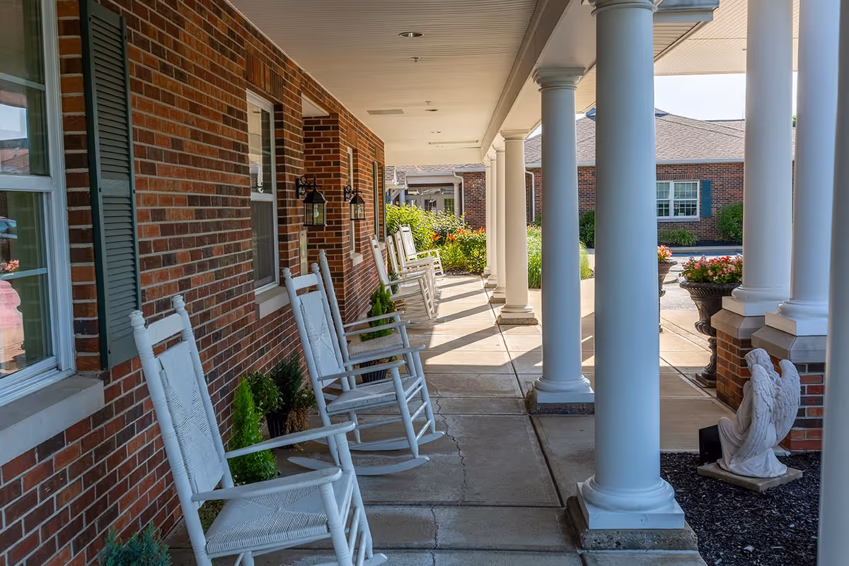 Covered front porch of a brick senior living facility with white rocking chairs, columns, potted plants, and a small angel statue.