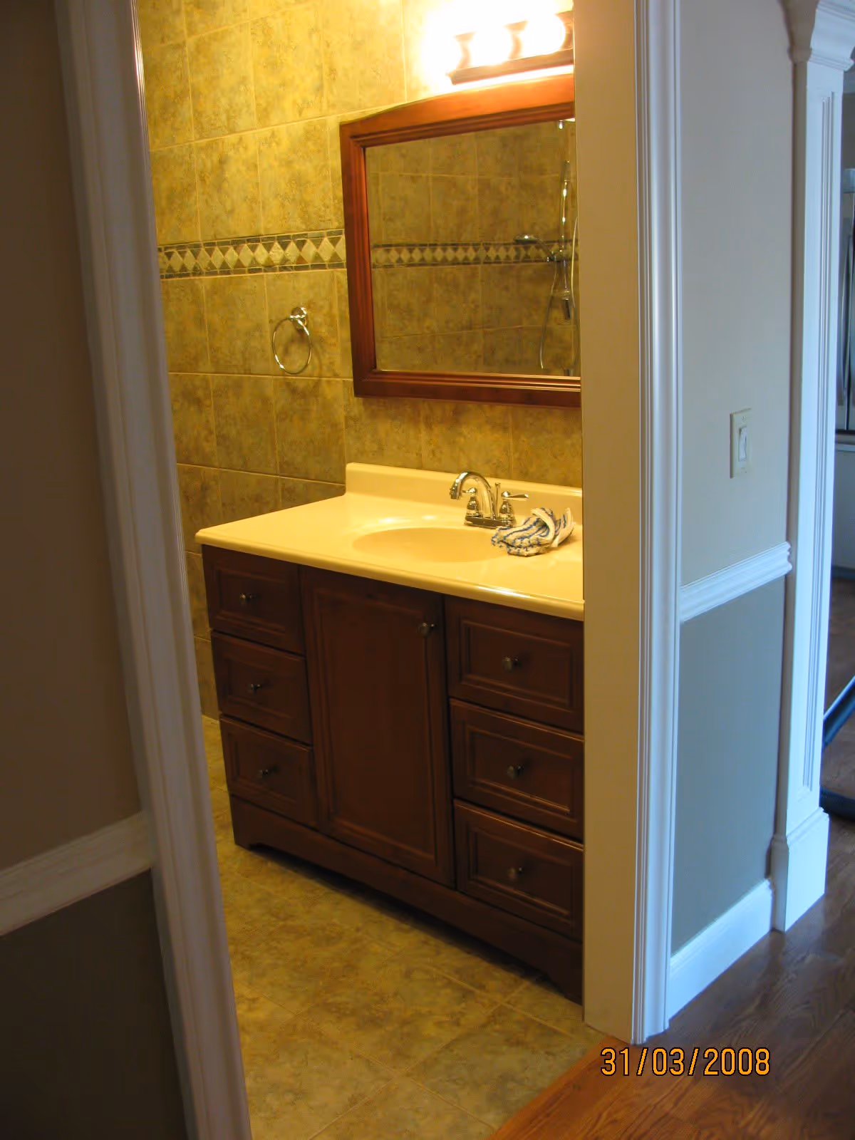 View into a bathroom showing a wooden vanity with a cream-colored countertop and built-in sink. Above the vanity is a rectangular mirror with a wooden frame, and a light fixture with three bulbs is mounted above the mirror. The walls and floor are tiled in a beige pattern with a decorative tile border on the wall. A towel ring is mounted on the wall to the left of the mirror.