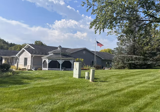 Exterior view of a senior living facility with a gazebo, flag, and green lawn.