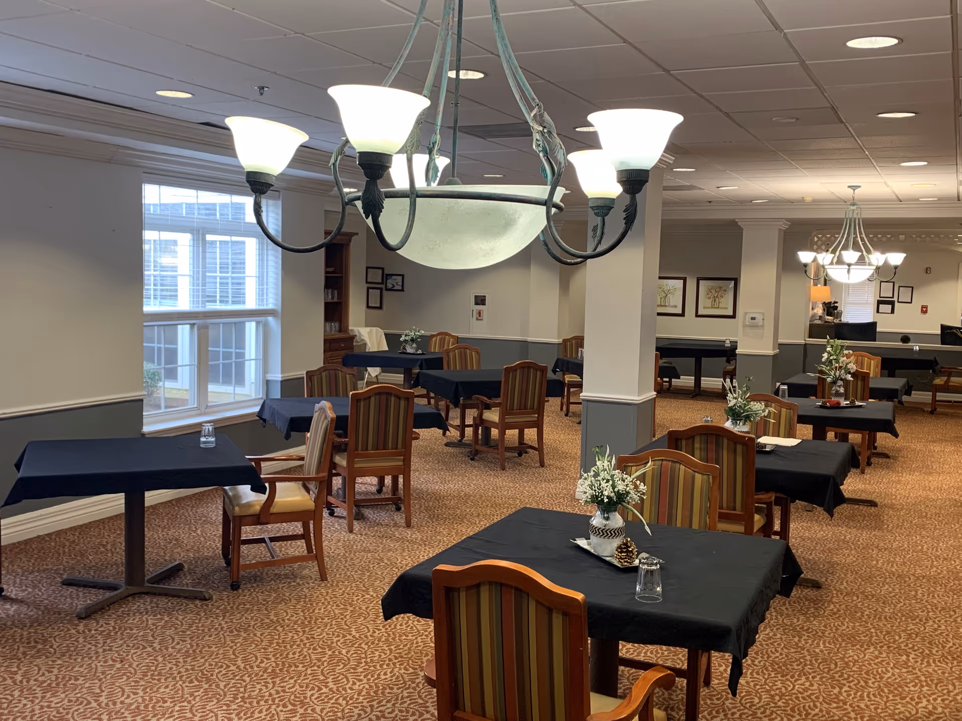 Spacious dining room with multiple tables covered in black tablecloths, wooden chairs, chandeliers, and floral centerpieces.