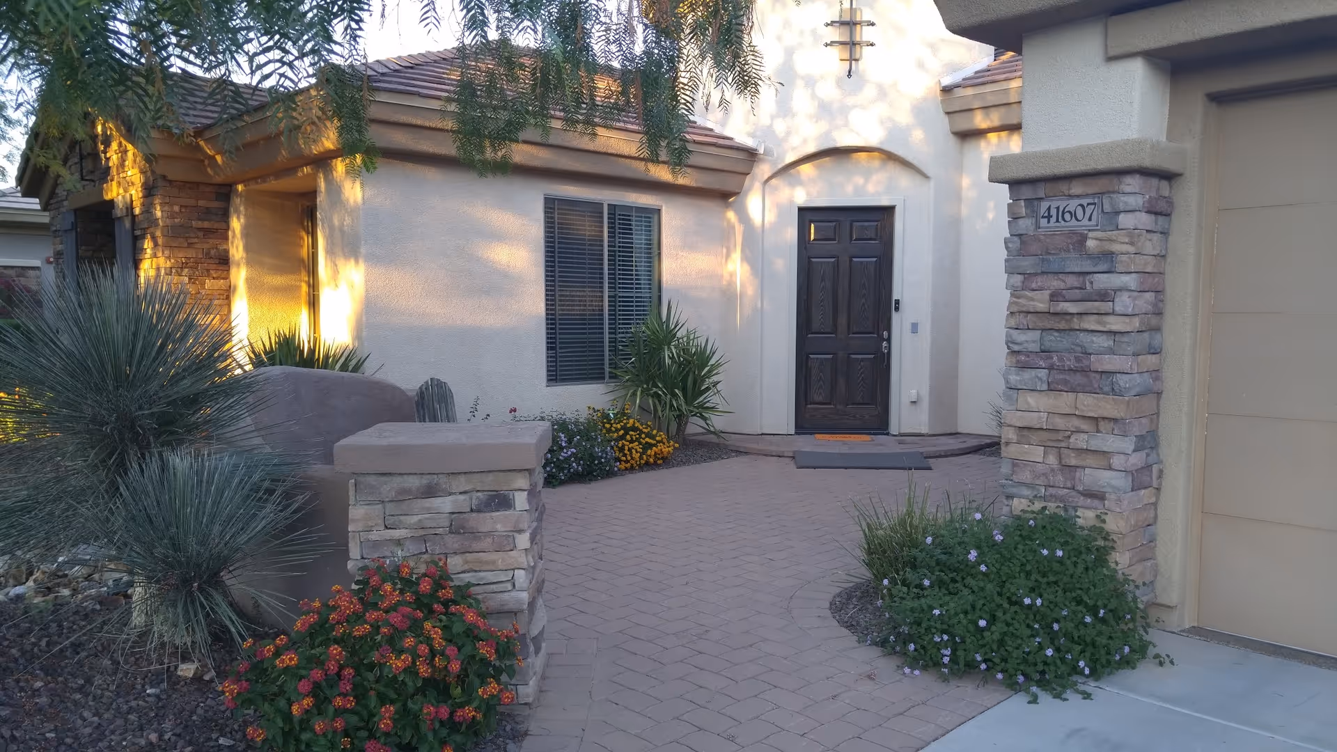 Front entrance of a single-story building with a dark wooden door, beige stucco walls, stone pillars, and a paved walkway surrounded by desert landscaping including various shrubs and flowering plants.
