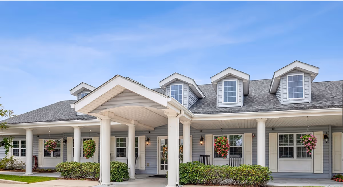Front entrance of a single-story senior living building with a covered portico, white columns, hanging flower baskets, and dormer windows.
