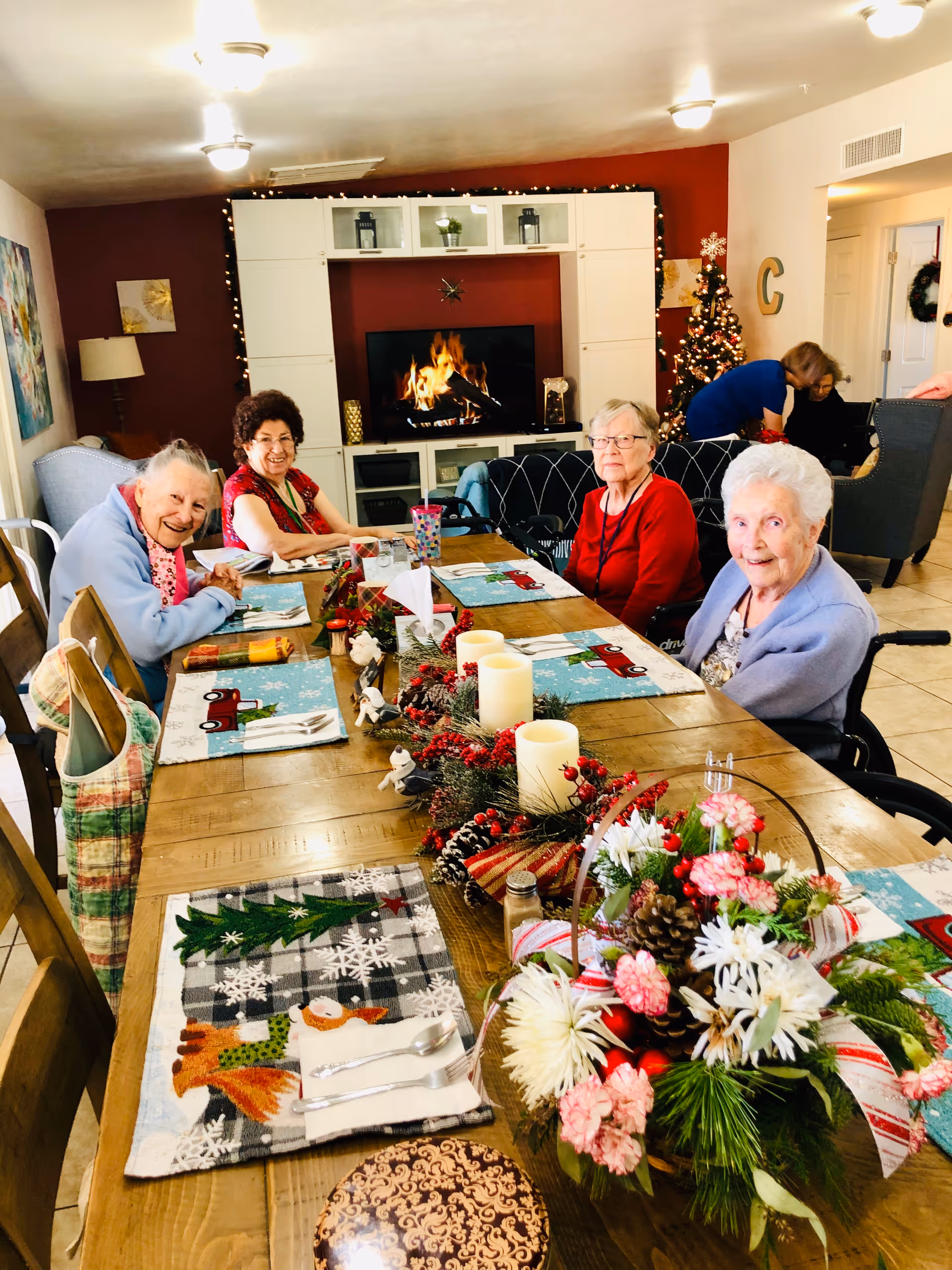 Four elderly women sitting around a wooden dining table decorated with festive holiday placemats, candles, and a floral centerpiece. In the background, there is a TV displaying a fireplace, a Christmas tree, and two other women engaged in an activity. The room has warm lighting and holiday decorations.
