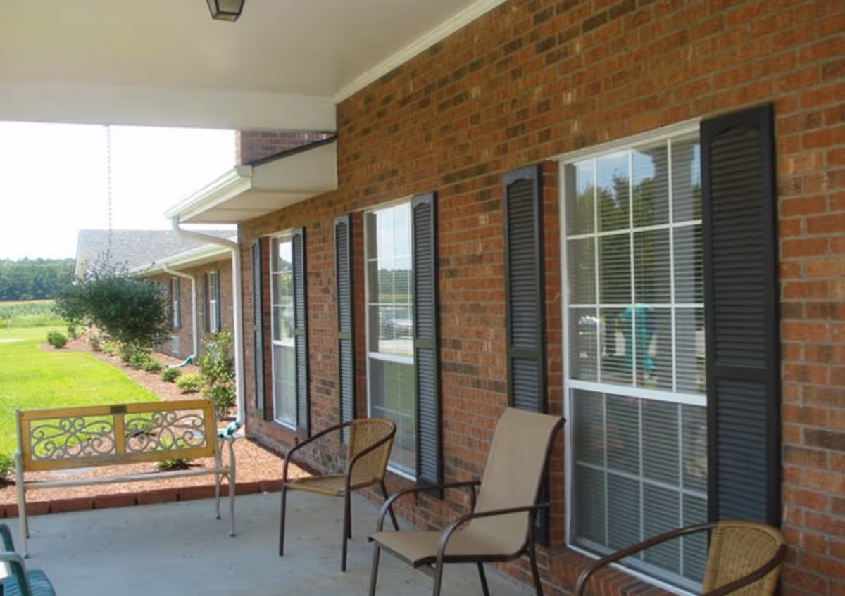 Covered outdoor patio area with a brick wall featuring multiple windows with black shutters. The patio has several chairs including a metal bench with decorative backrest, a wicker chair, and a beige sling chair. There is a well-maintained lawn and garden with shrubs and trees visible beyond the patio.