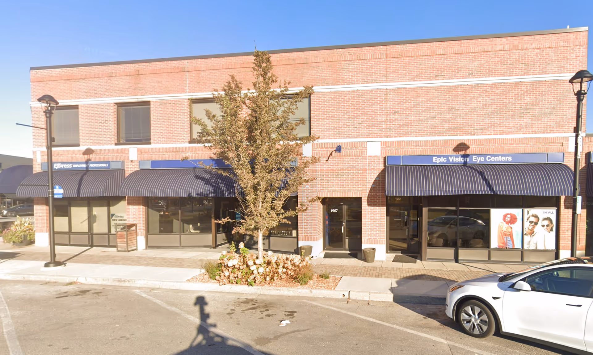 Exterior brick building with glass storefronts and navy awnings, sidewalk, tree, and a parked car.