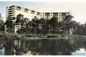 Multi-story building behind trees with a reflective body of water in the foreground.