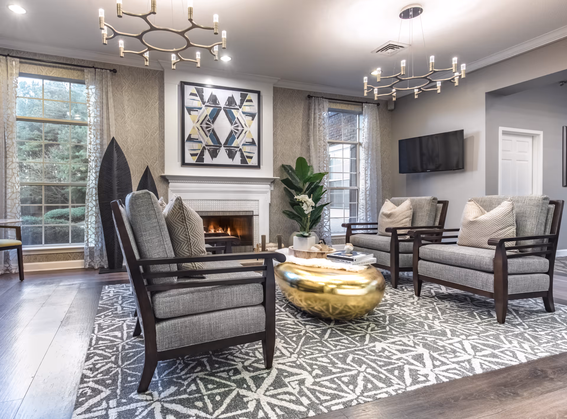 Well-appointed living room with a lit fireplace, four upholstered armchairs around a gold coffee table on a patterned rug, a wall-mounted TV, and large windows.