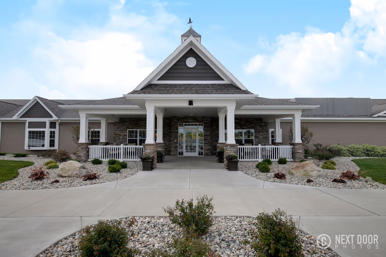 Front exterior view of Hathaway Hills Assisted Living & Memory Care building with a covered entrance, stone and siding facade, landscaped rock gardens, and a clear blue sky.