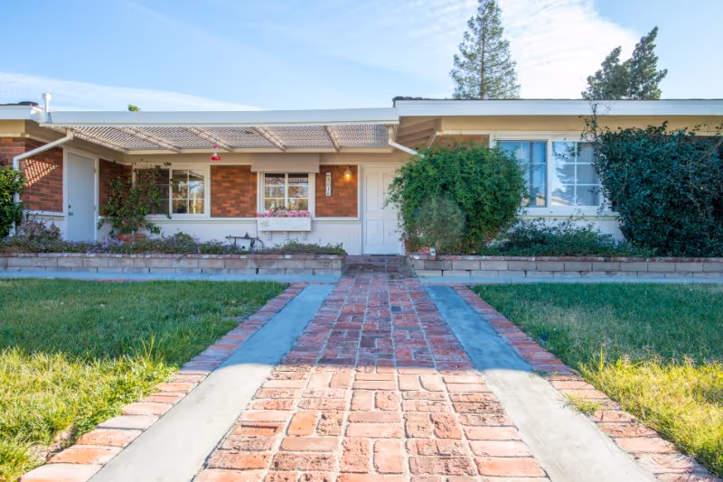 Brick walkway leading to the front entrance of a single-story building with windows, shrubs, and a covered porch.