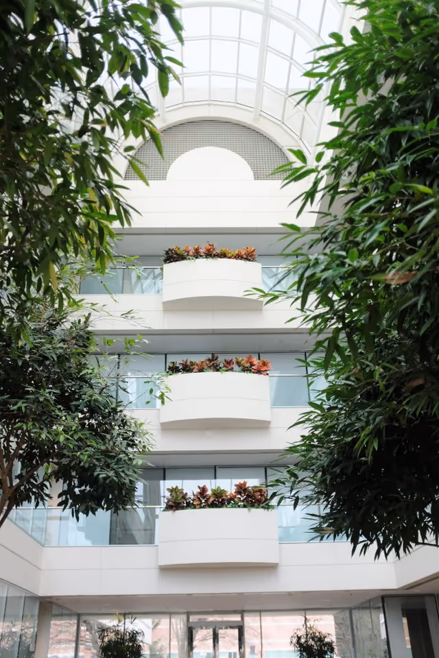 Indoor atrium with curved white balconies and lush green plants under a glass skylight.