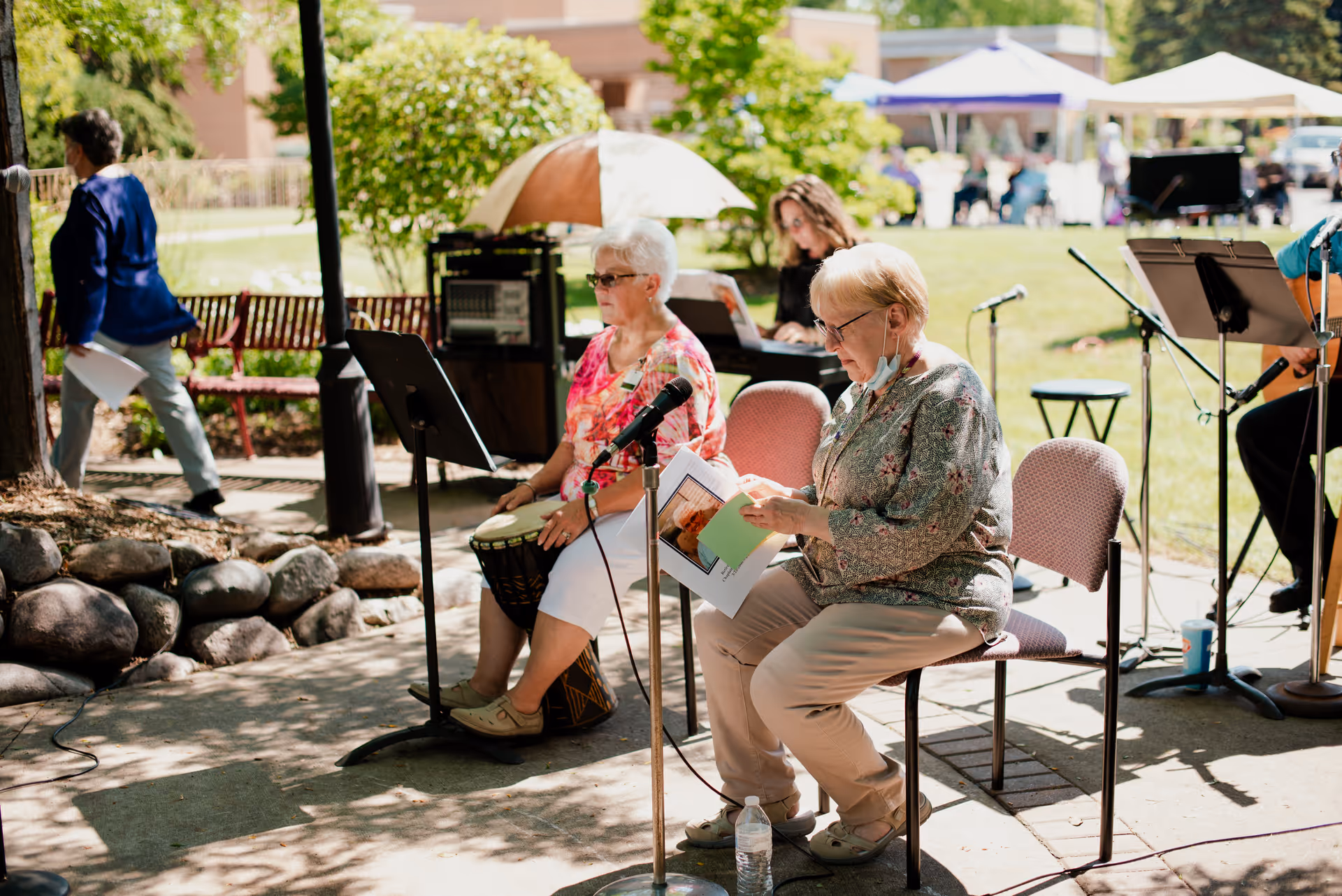 Two elderly women seated outdoors under a shaded area, one playing a drum and the other reading from a booklet with a microphone in front of her. In the background, a woman is playing a keyboard and other people are visible in a grassy area with tents and benches.