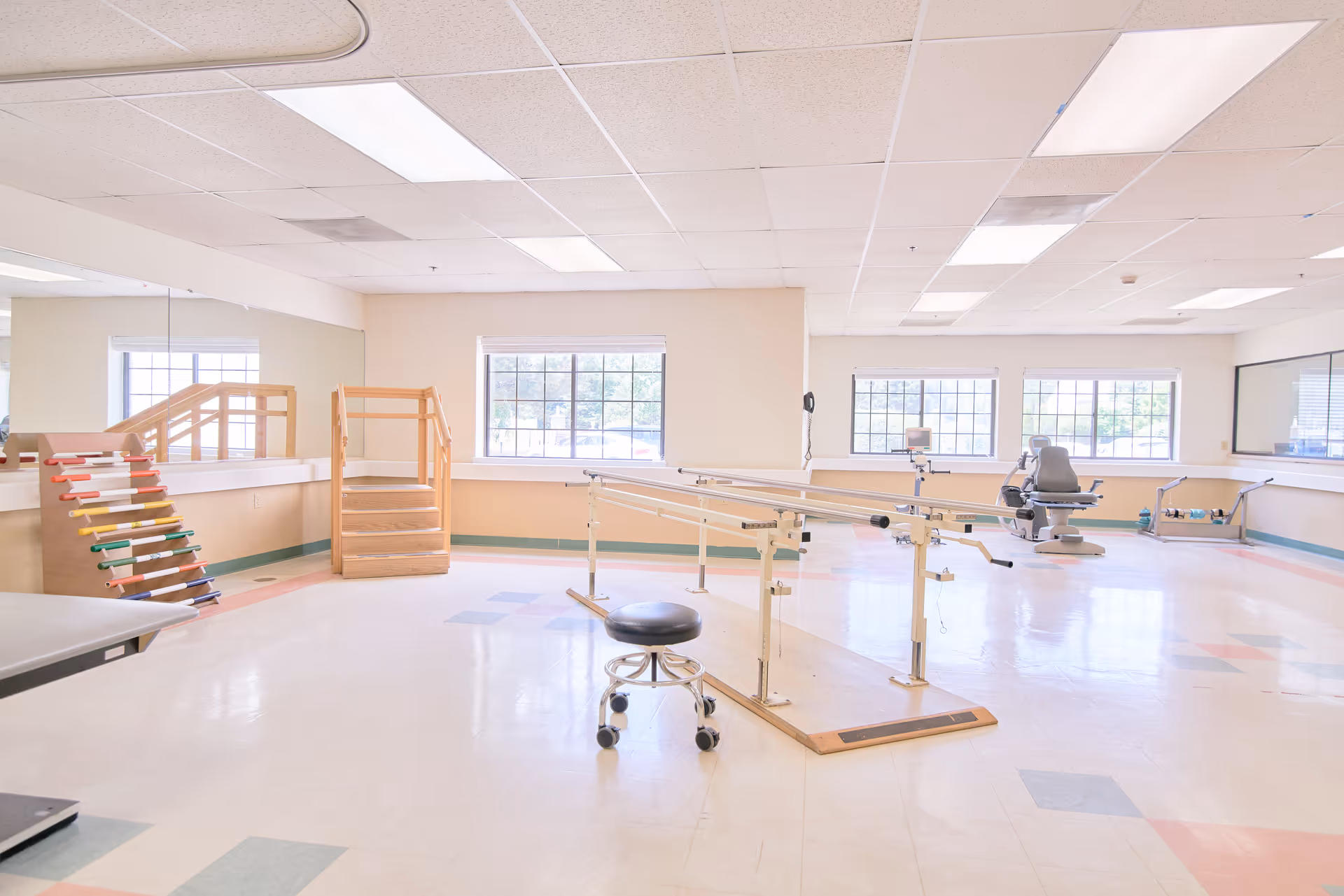 A bright and spacious physical therapy room with large windows letting in natural light. The room contains parallel bars for walking exercises, a small stool on wheels, a set of wooden stairs with handrails, a colorful ramp, and exercise equipment in the background. The floor has a light-colored tile with some colored square patterns.