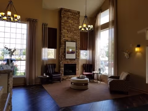 Warmly lit seating area with high windows, a stone fireplace, chandeliers, armchairs and a central ottoman.