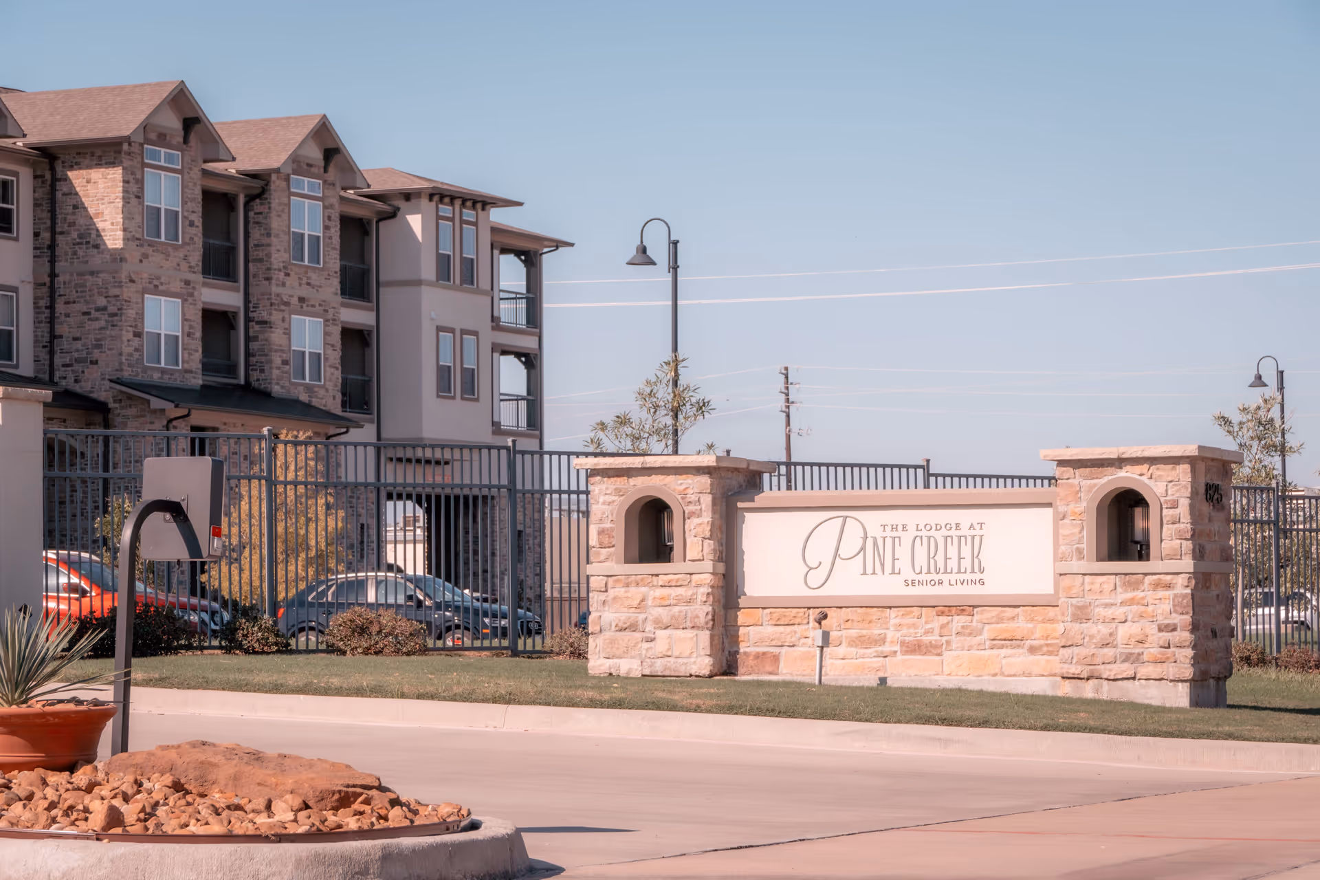 Exterior view of The Lodge at Pine Creek senior living facility showing a stone entrance sign with the facility name, a gated fence, a multi-story building with balconies, parked cars, and clear blue sky.