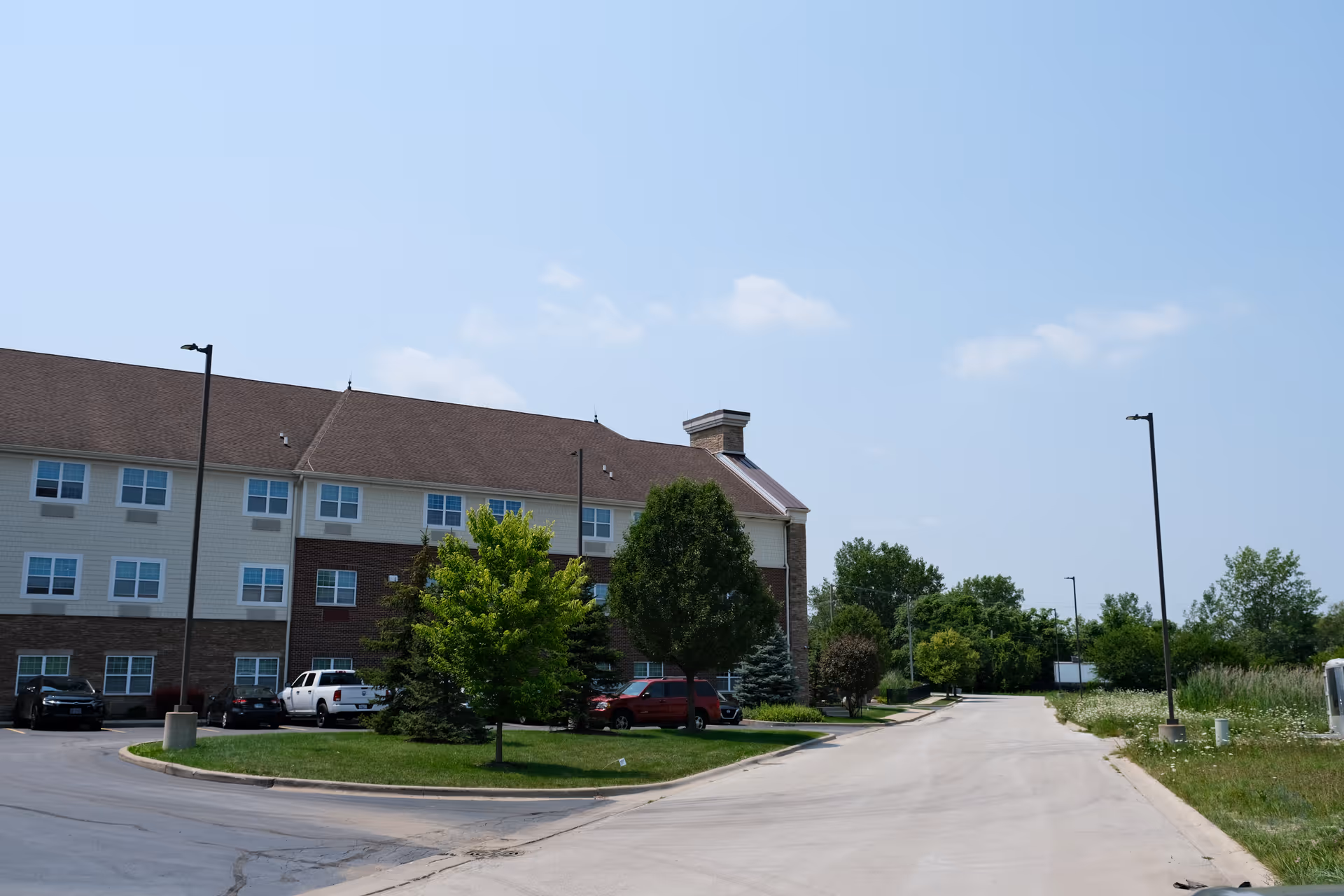 Three-story brick-and-siding residential building with parked cars, trees, and a driveway under a blue sky.