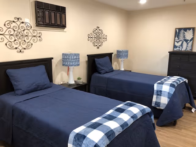 A bedroom with two twin beds covered in dark blue bedspreads and blue and white checkered blankets folded at the foot. Each bed has a matching blue pillow. Between the beds is a small nightstand with a white lamp featuring a blue patterned lampshade and a small plant. The walls are beige with decorative metal wall art above each bed. A dark wooden dresser with a framed botanical print on top is visible in the corner.
