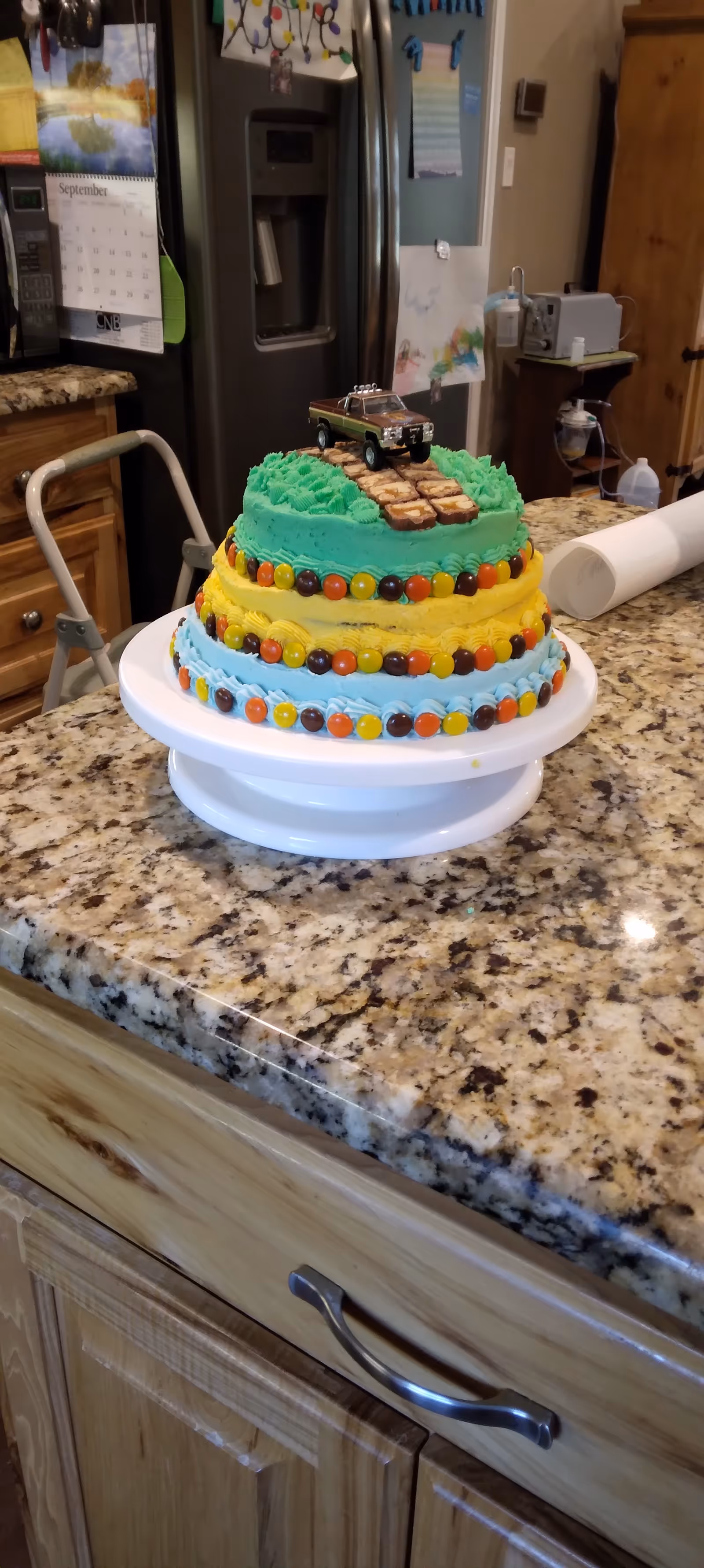 Colorful multi-tier cake with a toy car topper sits on a kitchen counter.