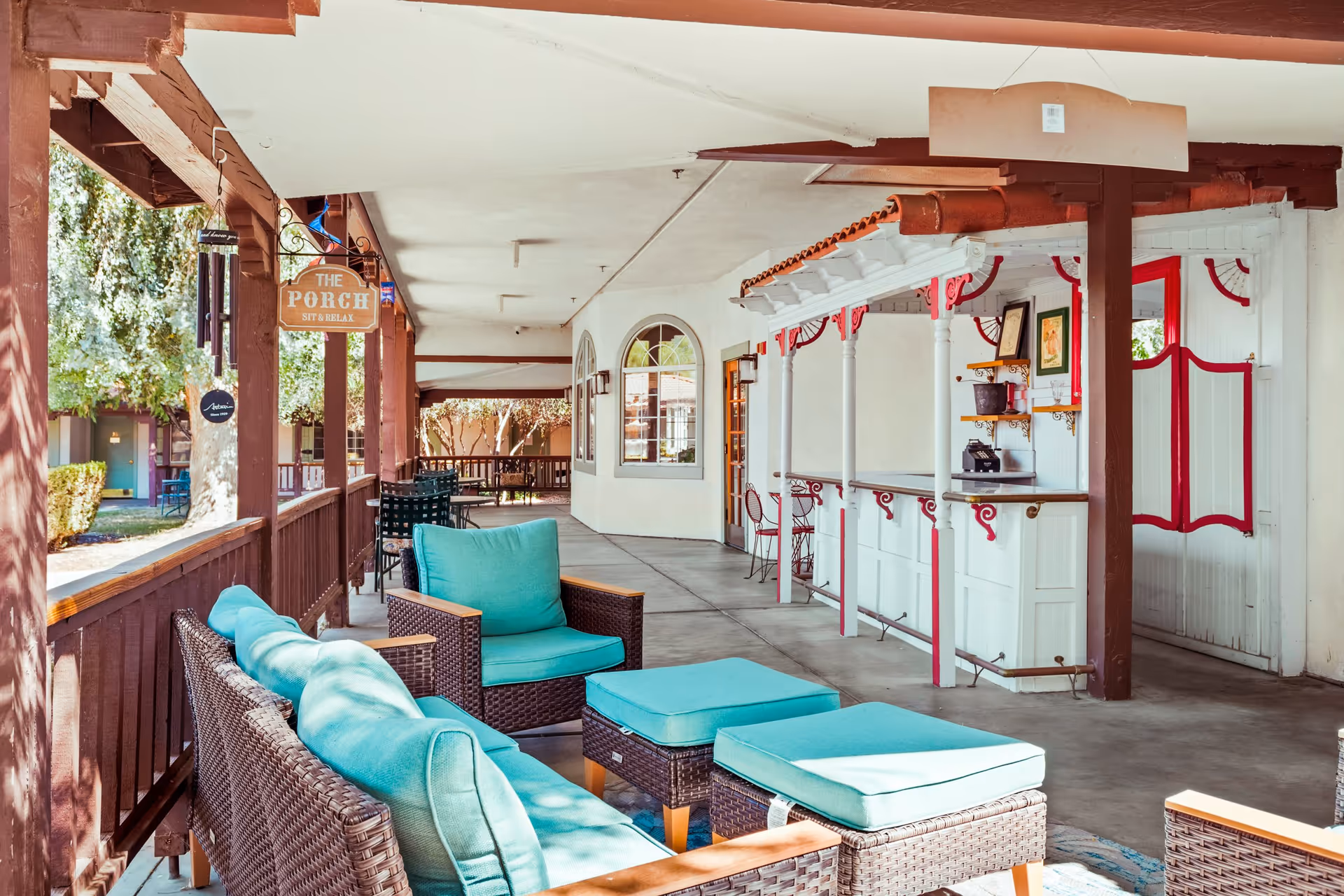 A covered outdoor porch area with wicker furniture featuring turquoise cushions, a small bar with white and red trim, and a sign that reads 'The Porch Sit & Relax'. Trees and greenery are visible beyond the porch railing.