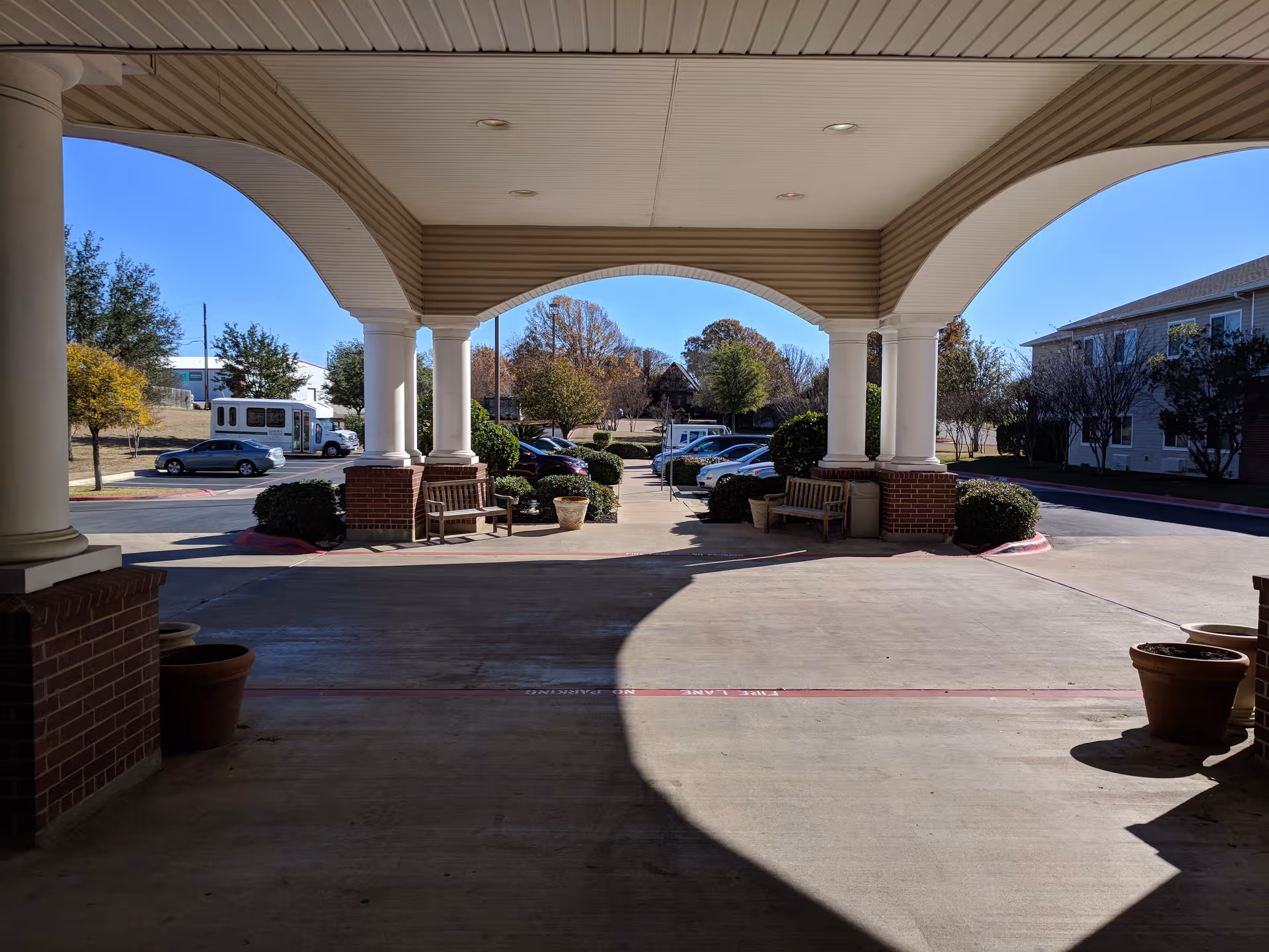 Covered outdoor entrance with tall white columns, benches, planters, and parked cars nearby.