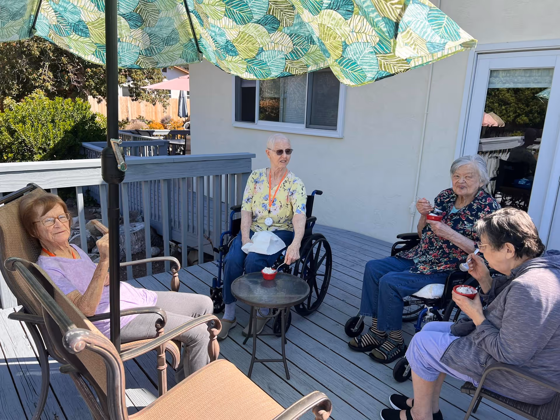 Four elderly residents sit on a shaded outdoor deck, enjoying drinks together under a large umbrella.