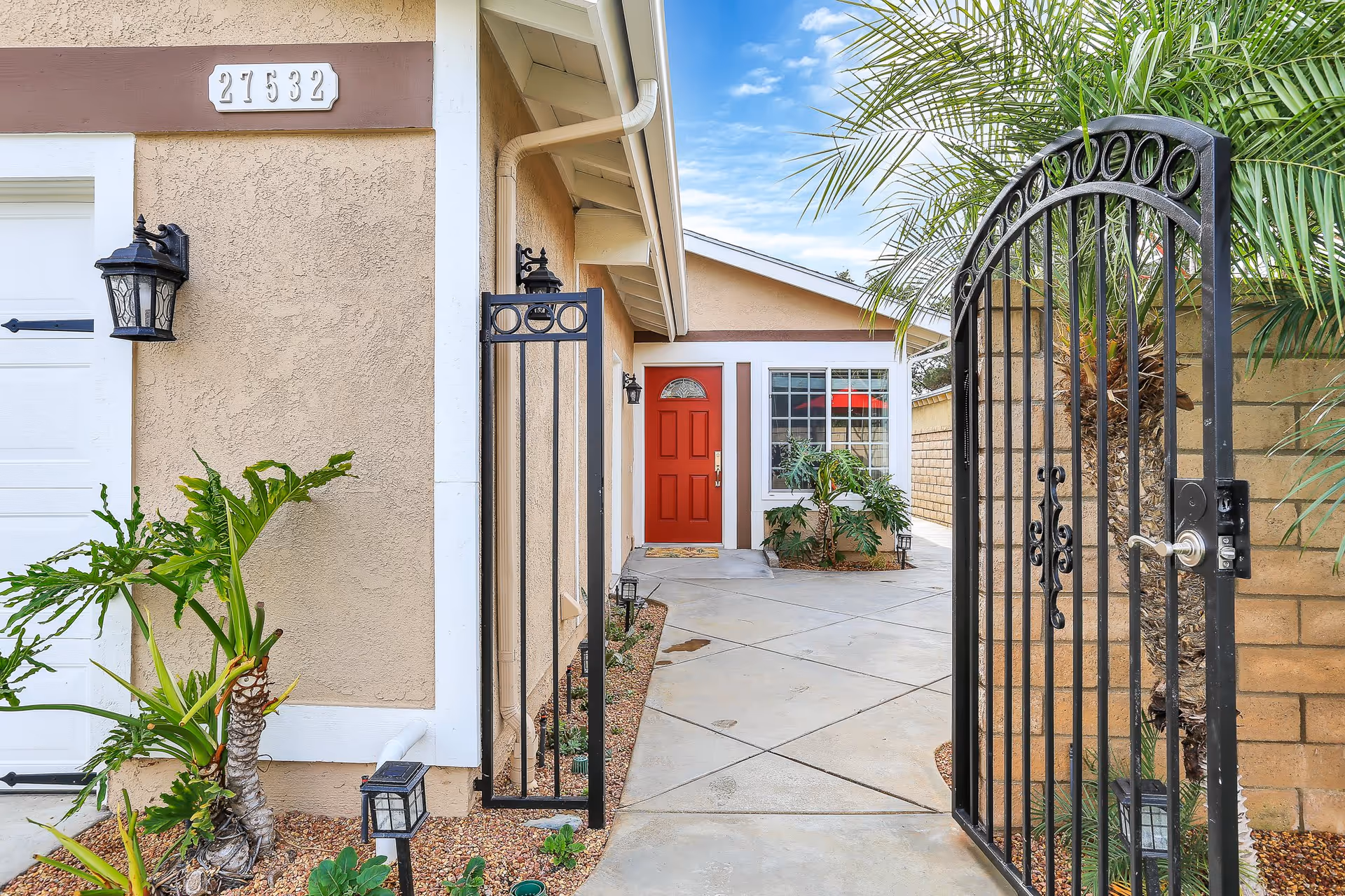 Wrought-iron gate opening to a paved courtyard leading to a beige stucco house with a red front door and plants.