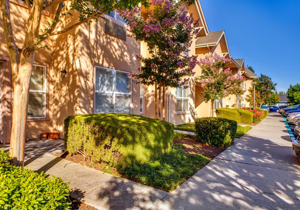 Sunny front exterior of a row of beige townhome-style units with manicured hedges, flowering trees, and a sidewalk beside parked cars.