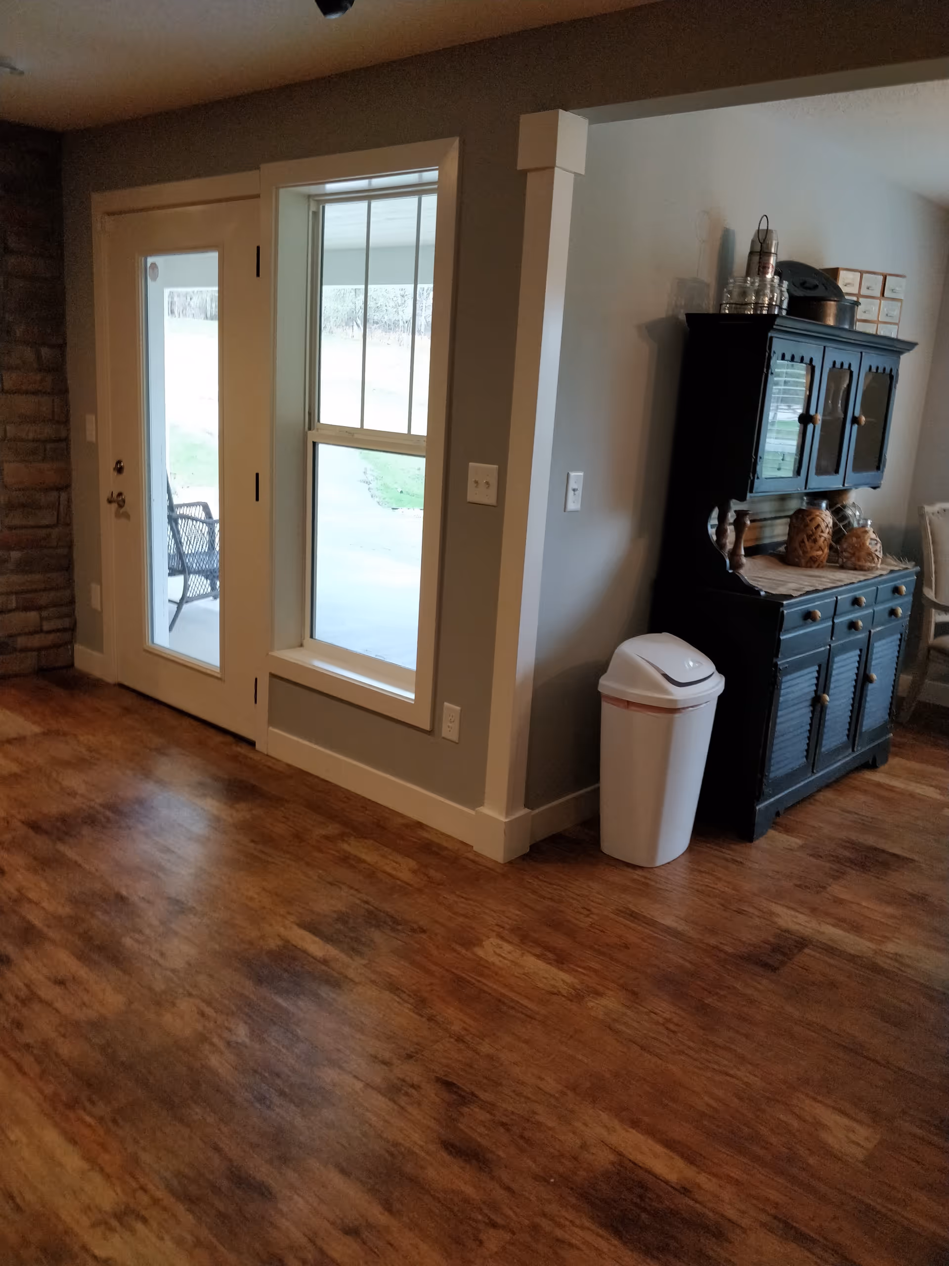 Interior living area with wooden floor, glass door, window, trash can, and dark cabinet.