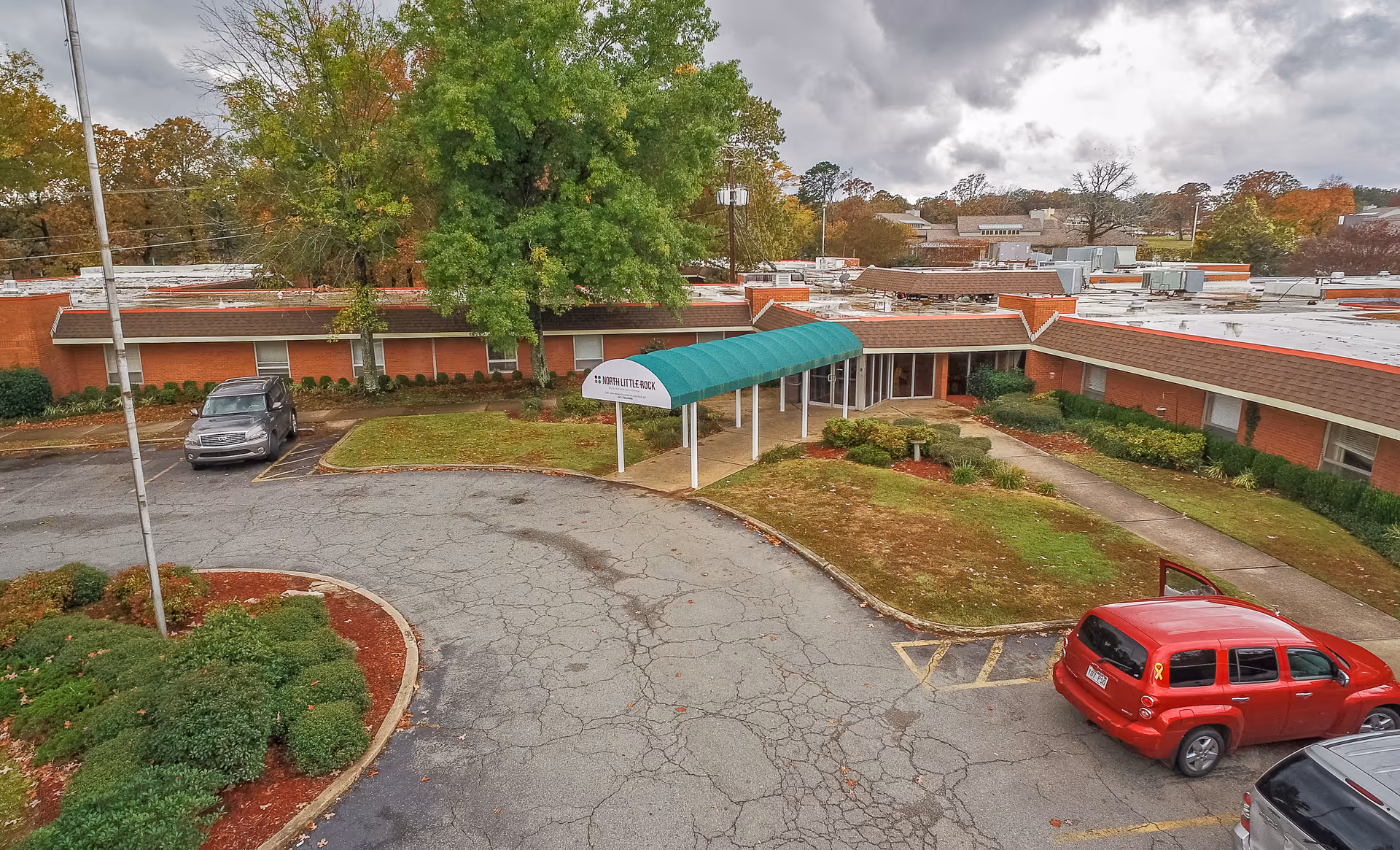 Exterior view of The Blossoms at North Little Rock Rehab & Nursing Center showing a single-story brick building with a green awning over the entrance. There are a few cars parked in the cracked parking lot, and trees with autumn foliage surround the area under a cloudy sky.