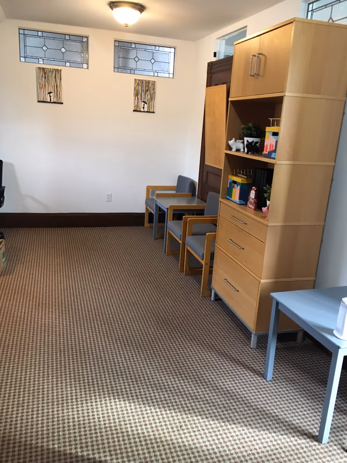 Interior senior-living room with beige checkered carpet, chairs, cabinet, and wall artworks.