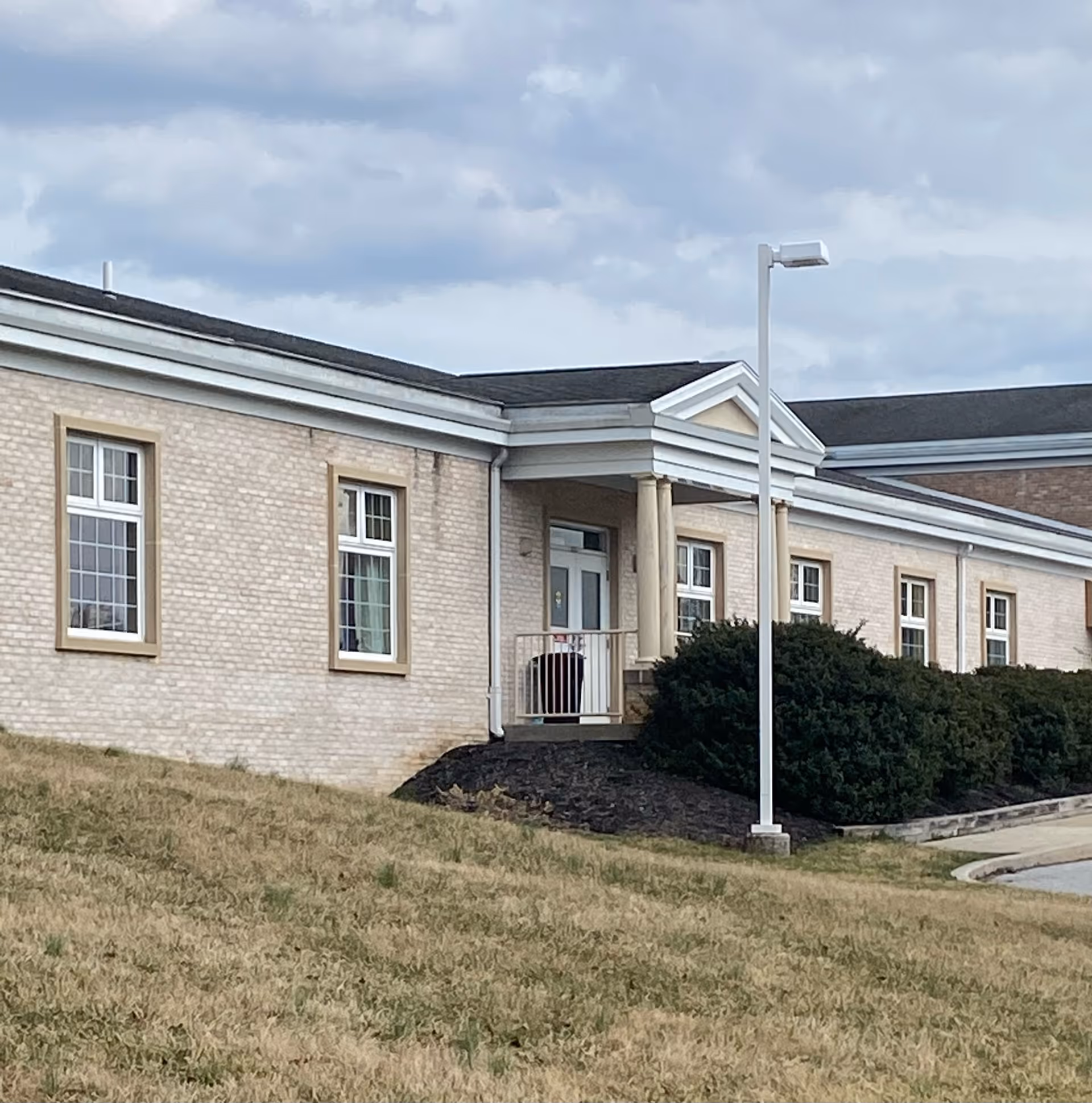 Exterior view of a single-story brick building with multiple windows and a small entrance porch supported by columns. There is a grassy area in the foreground and a cloudy sky above.