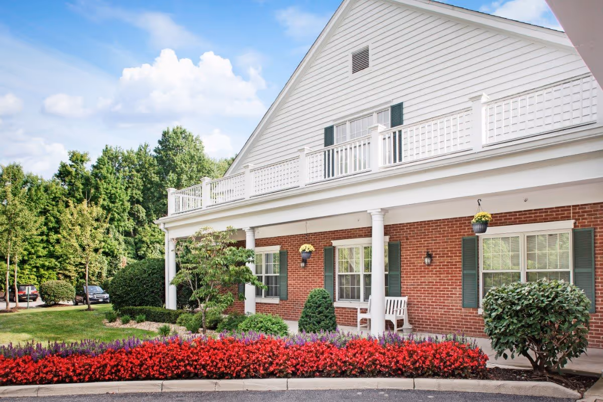 Exterior view of a senior living facility with a brick facade, white columns, and a white balcony railing. There are green shutters on the windows, a white bench on the porch, and well-maintained landscaping including red and purple flowers, bushes, and trees under a partly cloudy blue sky.
