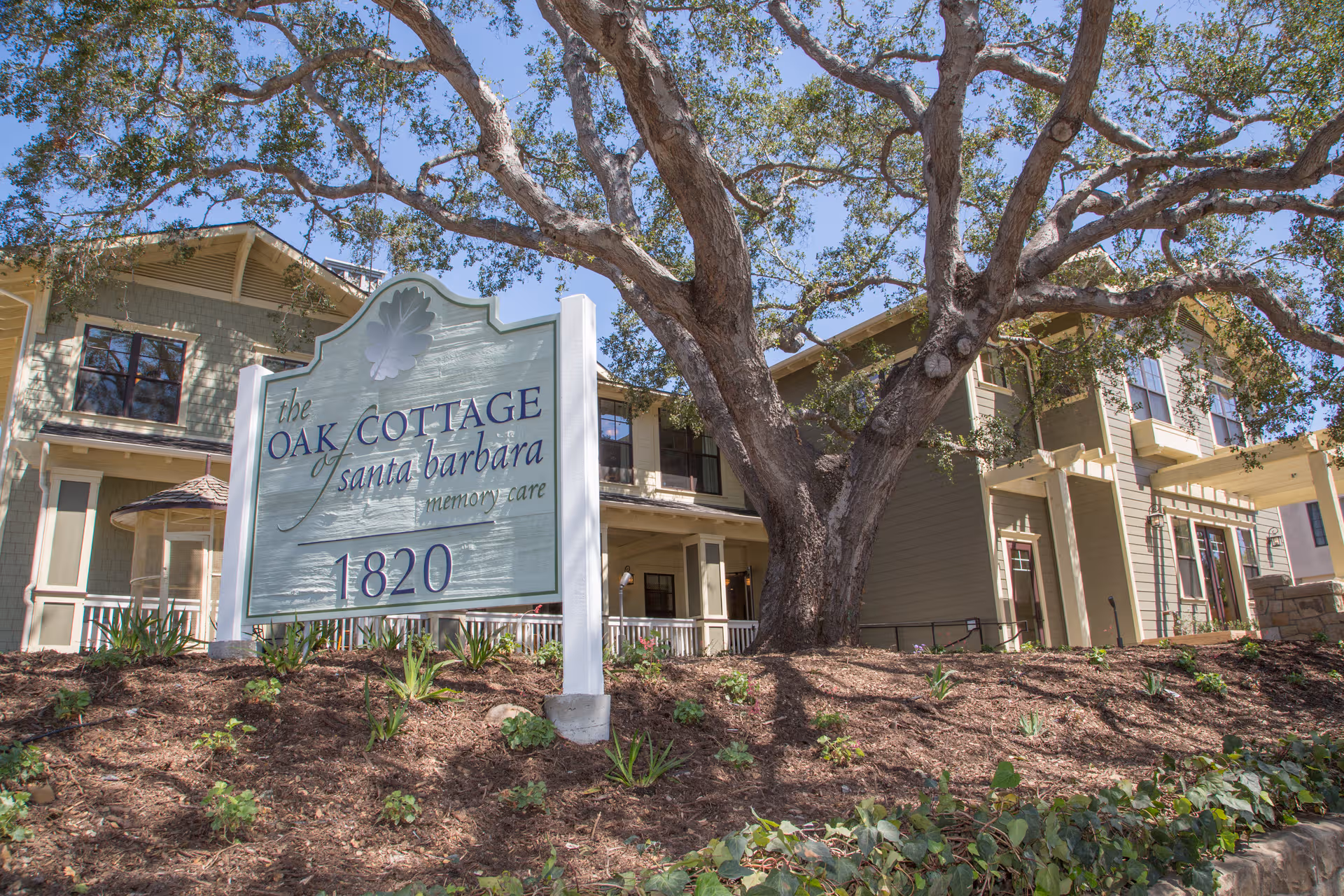 Exterior view of Oak Cottage of Santa Barbara Memory Care with a large sign and tree.