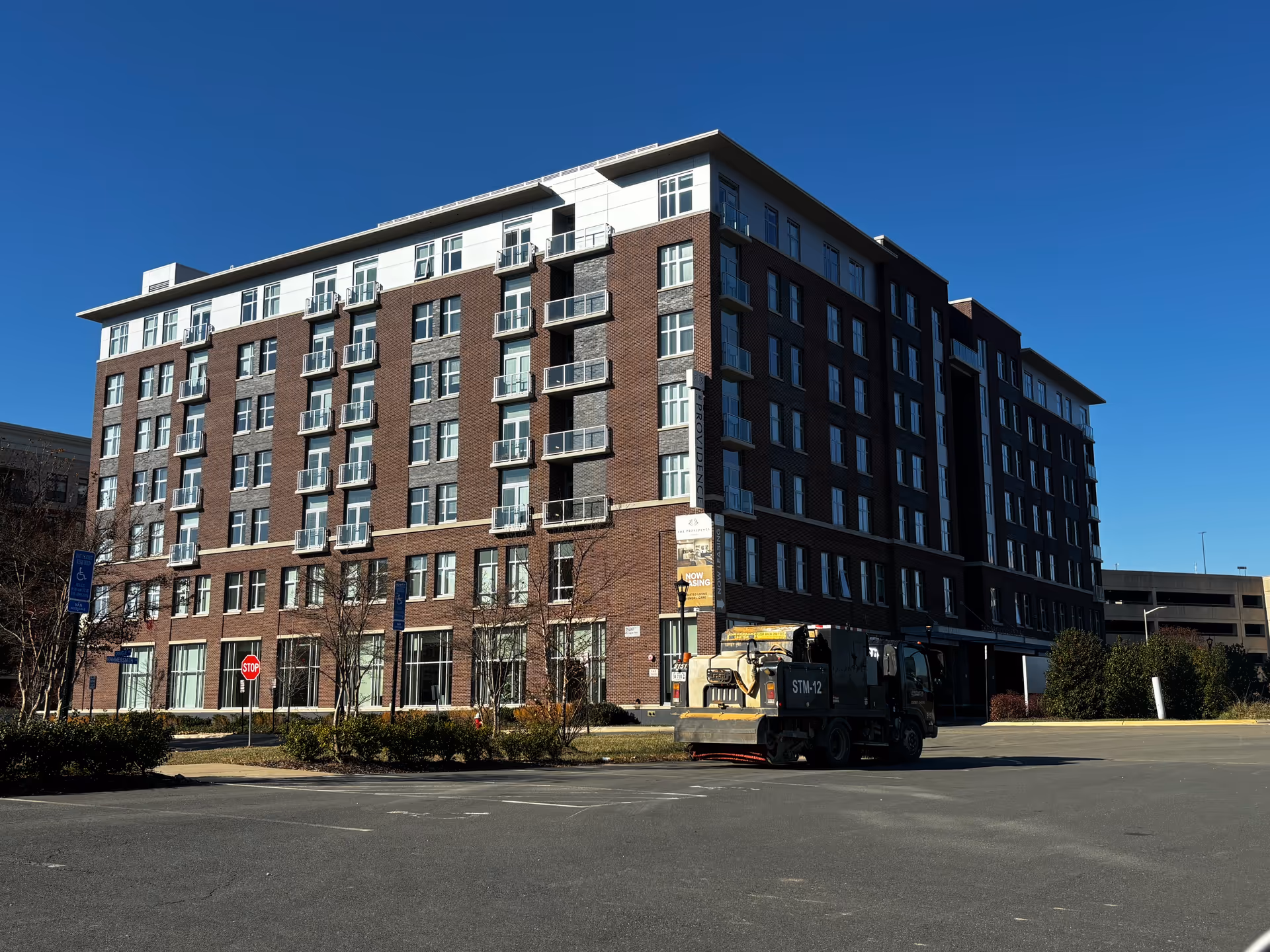 Exterior view of a multi-story brick and glass building under a clear blue sky, with a street sweeper vehicle parked in the foreground and some small trees and bushes around the building.