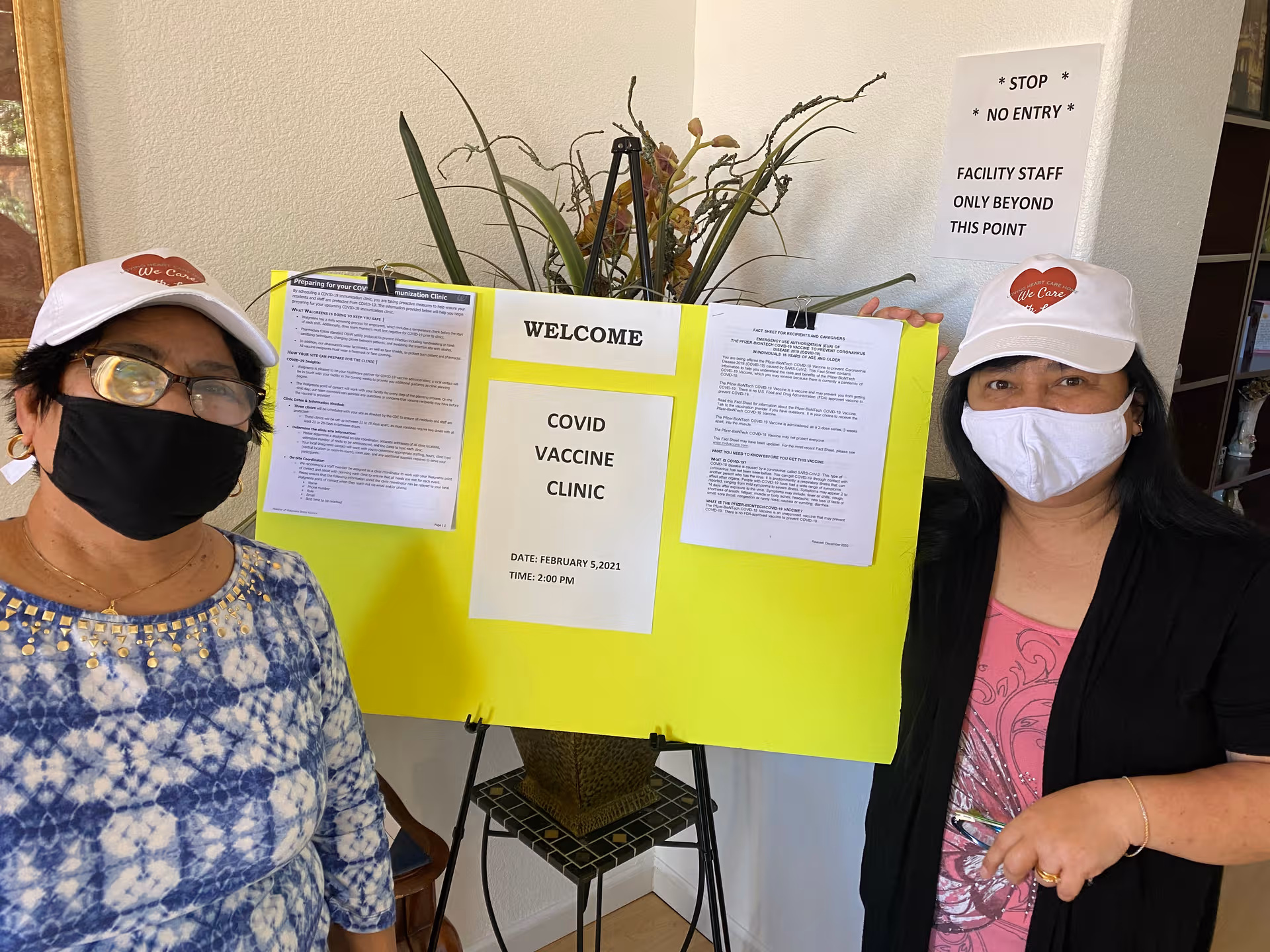 Two women in masks stand beside a bright yellow COVID vaccine clinic sign.