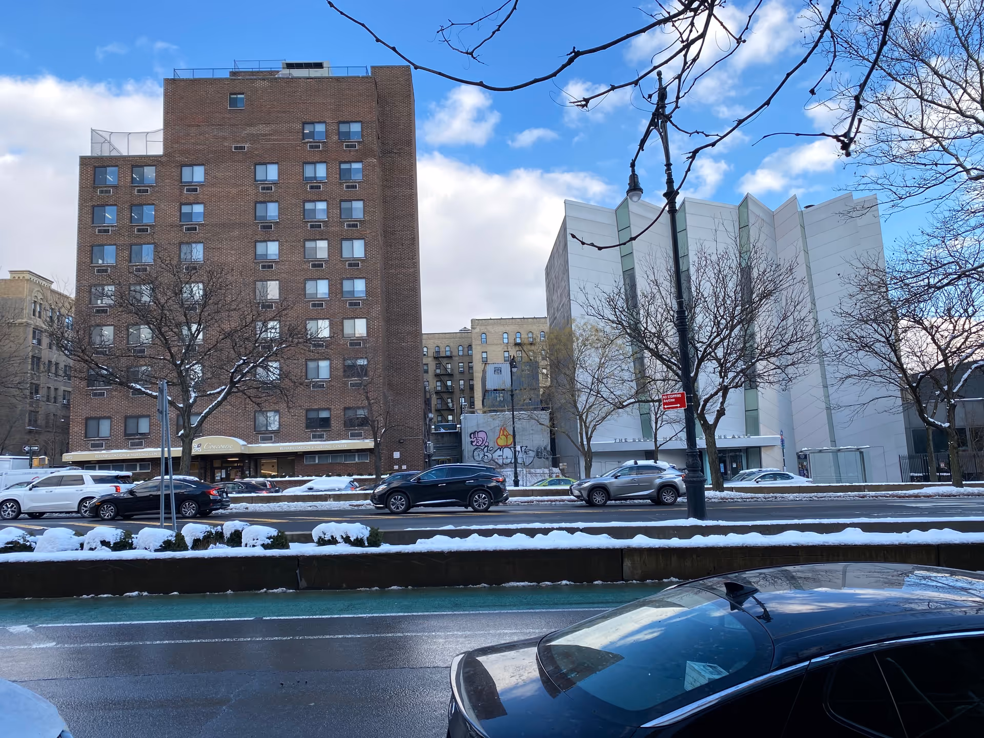 Snowy urban street exterior with brick and white buildings, trees, and parked cars.
