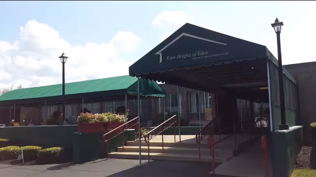 Entrance of Eden Heights Of Eden facility with a covered walkway and ramp, flanked by two lamp posts and surrounded by greenery under a partly cloudy sky.